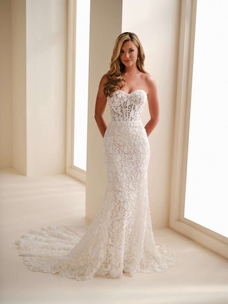Full-length front view of the E621 ivory and almond fit-and-flare lace bridal gown beside a window