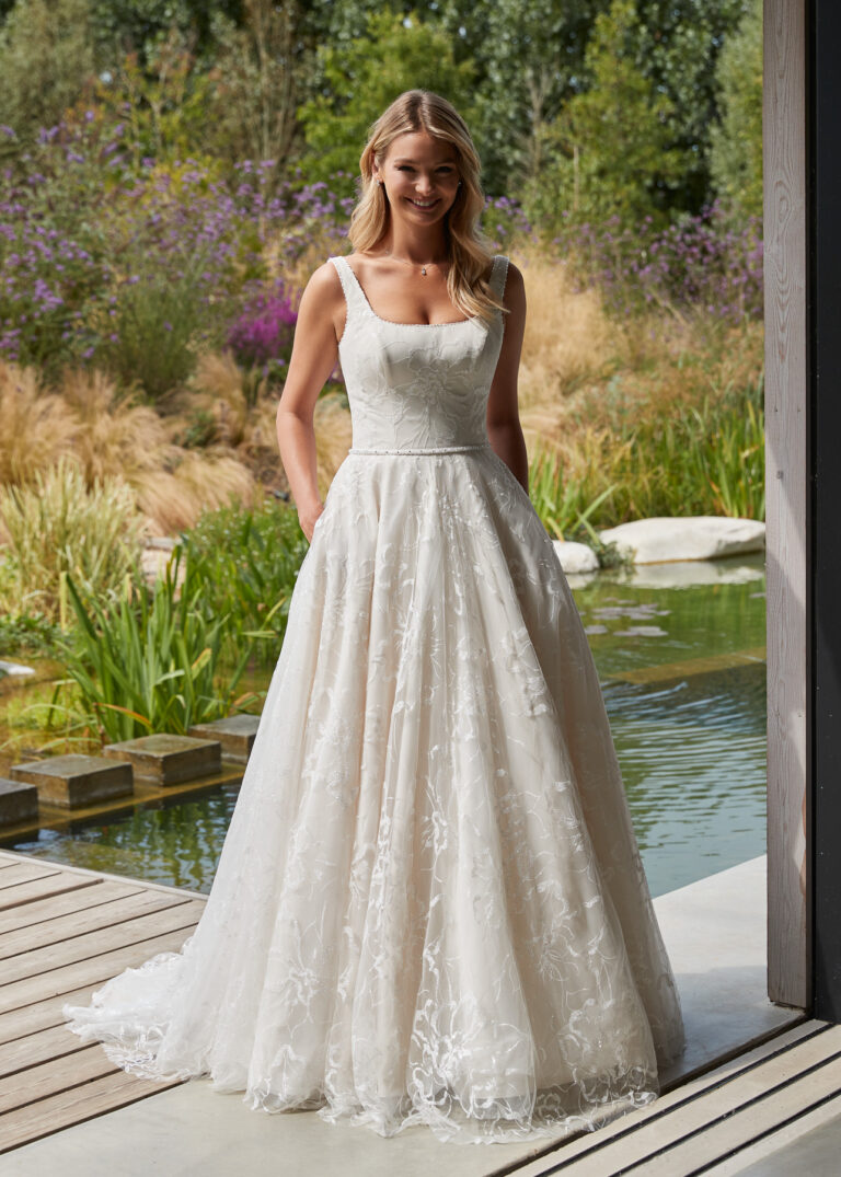Full-length front view of the Lyanna ivory and silver A-line bridal gown with square neckline beside a garden pond