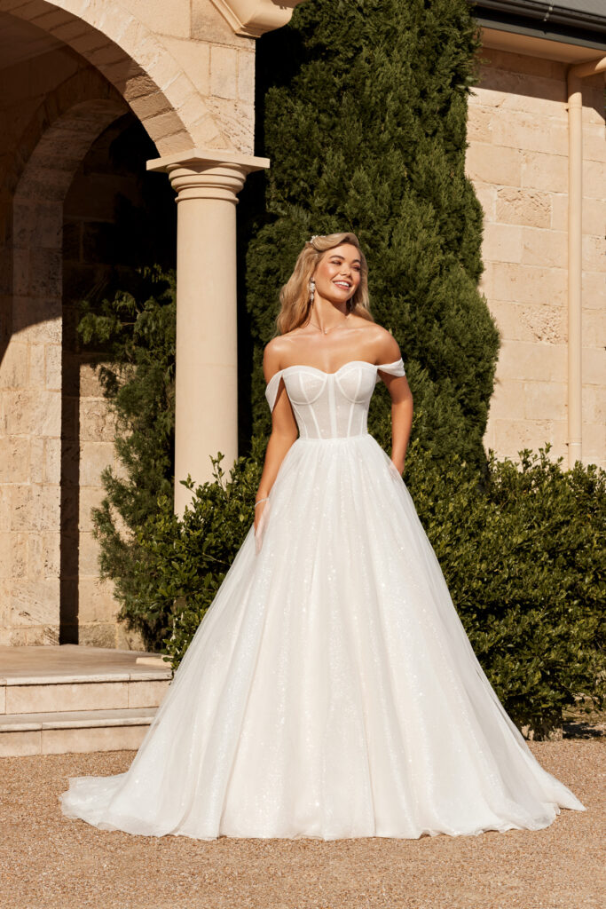 Full-length front view of the Lazaris Y3103 ivory ball gown with off-the-shoulder corset bodice outdoors