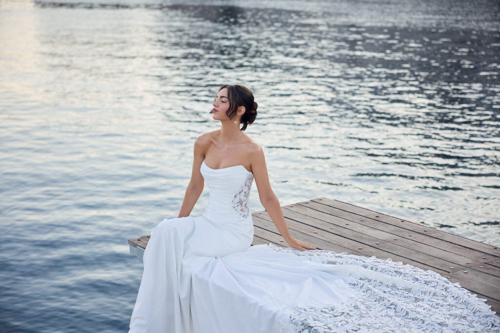 Seated side view of the Lucie bridal gown with the lace train spread across a waterfront jetty