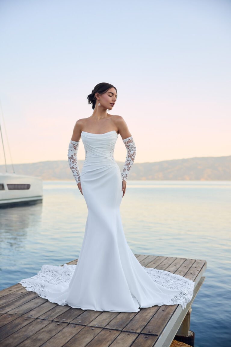 Full-length front view of the Lucie Y3180 ivory fit-and-flare gown with detachable lace sleeves beside the water
