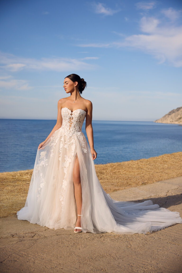 Full-length front view of the Betty Y3190FI ivory ball gown with floral bodice, slit skirt and train beside the sea